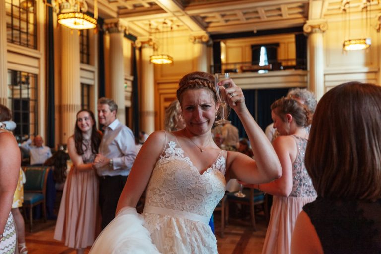 Bride in a white lace dress holding a glass, surrounded by guests in the council house ballroom with chandeliers and columns.