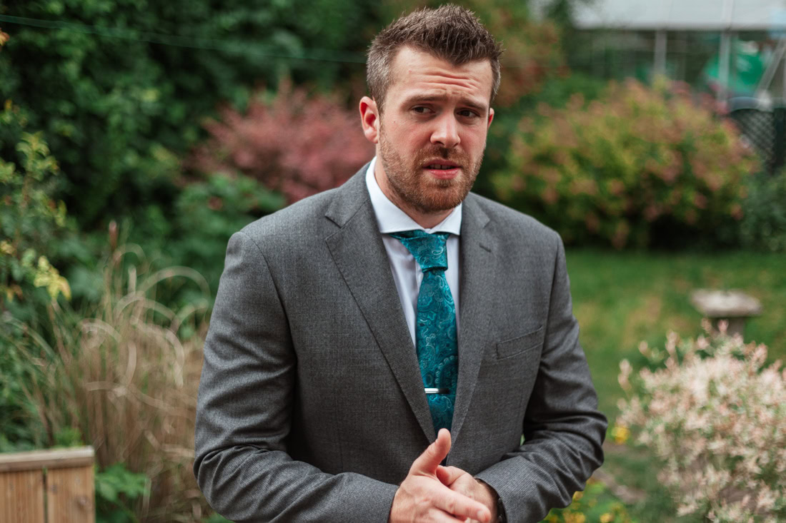 A groom in a grey suit with a teal tie stands in a garden with greenery and flowers in the background.