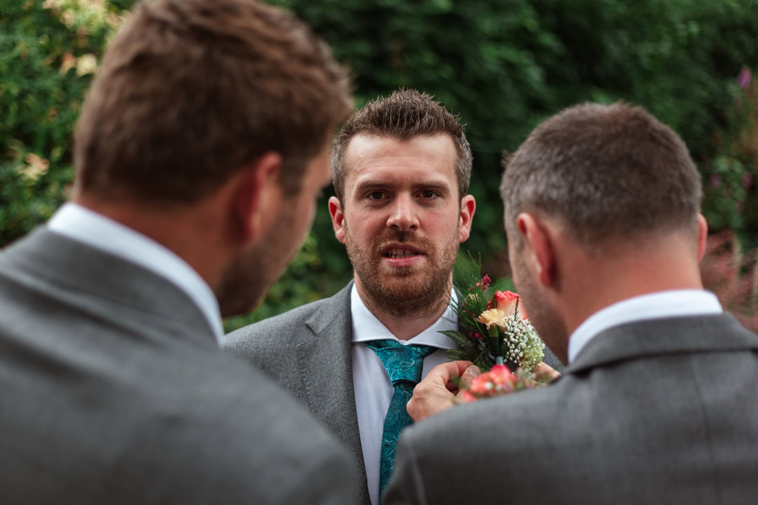 Three men in suits stand together as one adjusts the groom's boutonniere; teal tie and flowers in the background.