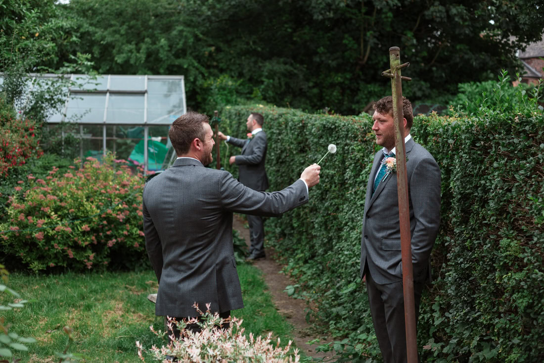 Three men in suits stand in a garden; one playfully holds a dandelion puff toward another.