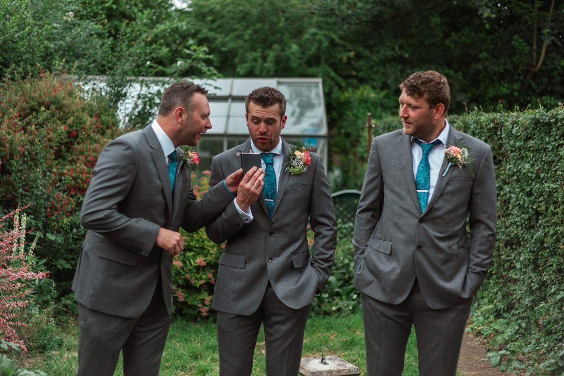 Three men in grey suits with teal ties stand in a garden while drinking out of a hip flask.