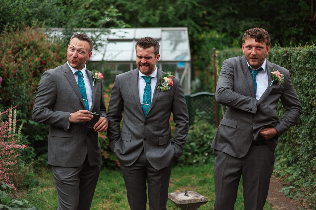 Three men in grey suits with teal ties stand in a garden with a greenhouse behind them.