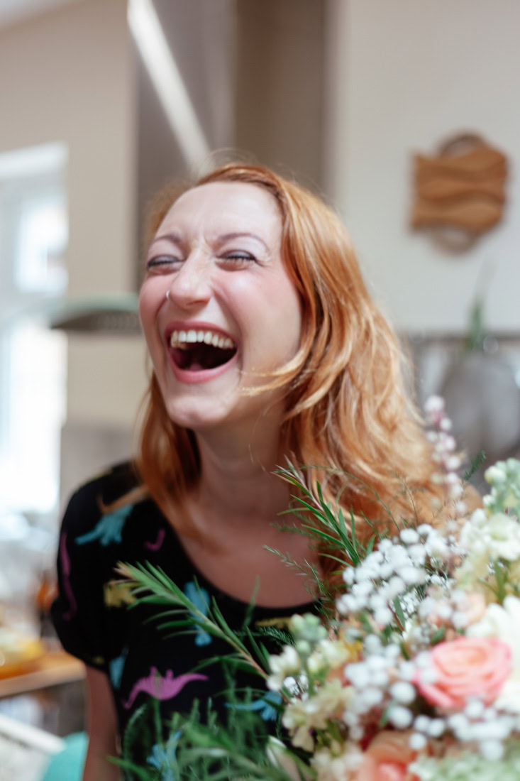 Person with long wavy hair in a dinosaur-print shirt holds a bouquet of flowers indoors laughing.