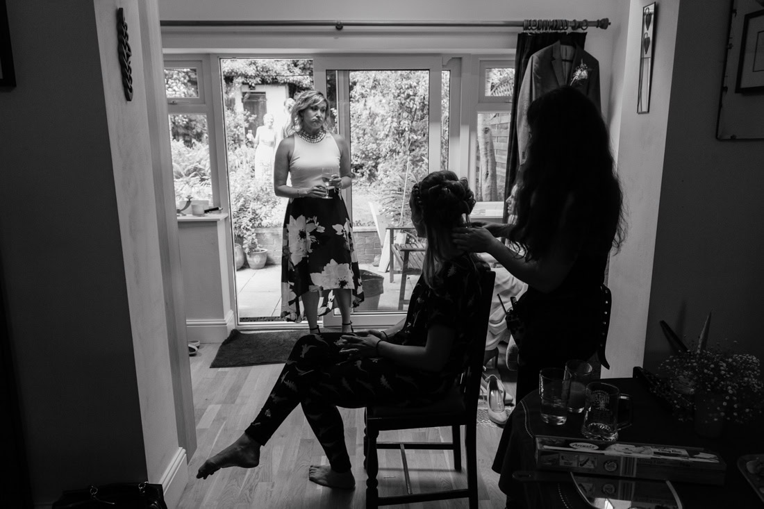 One person styles another’s hair indoors while someone stands in the doorway holding a drink, garden visible outside.