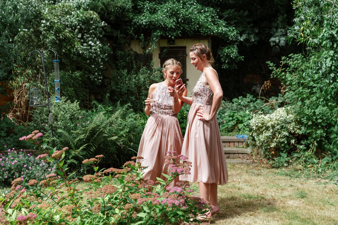 Two bridesmaids in pink dresses stand in a garden surrounded by greenery and flowers, talking together.