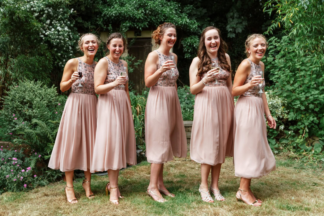 Five bridesmaids in matching pink dresses hold drinks while posing in a garden with greenery and flowers.