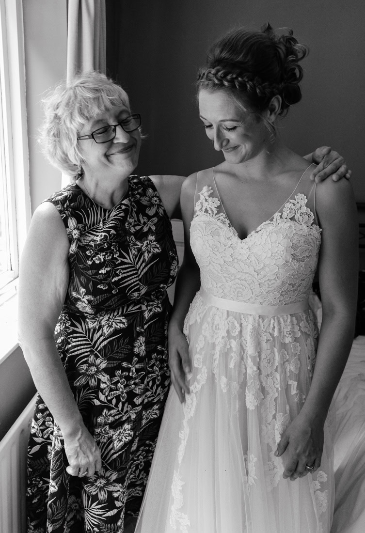 Two people stand together smiling, one in a floral dress and the other in a lace wedding gown.