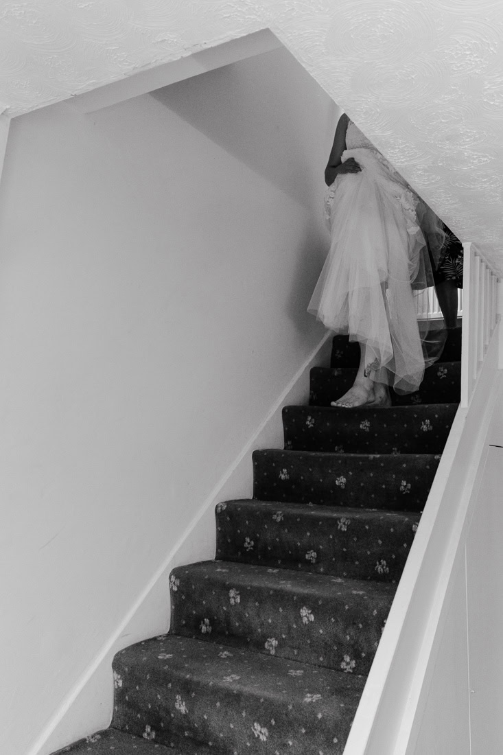 A person in a wedding dress walks up a floral-patterned carpeted staircase; black and white photo.