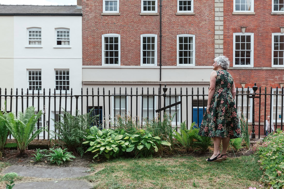 Person in a floral dress stands in a garden, facing a black fence and brick buildings with many windows.