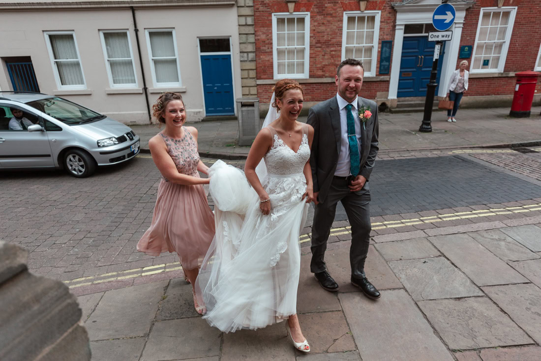 Bride walks with a man in a suit and a woman in a pink dress along a cobblestone street.