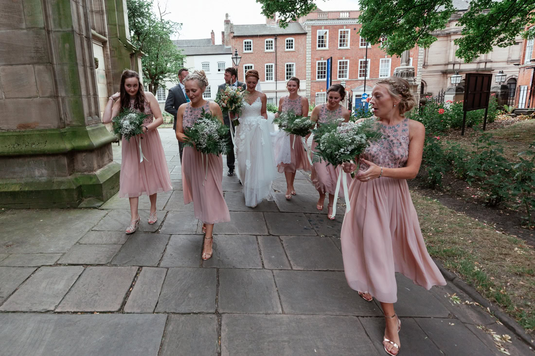Bride and bridesmaids in pink dresses walk on a stone path outside St Marys church, holding bouquets.