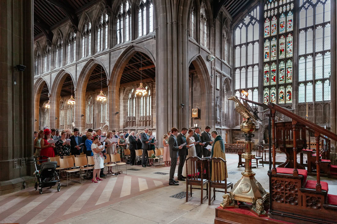People stand and sing inside a gothic-style church with tall stained glass windows and ornate architectural details.