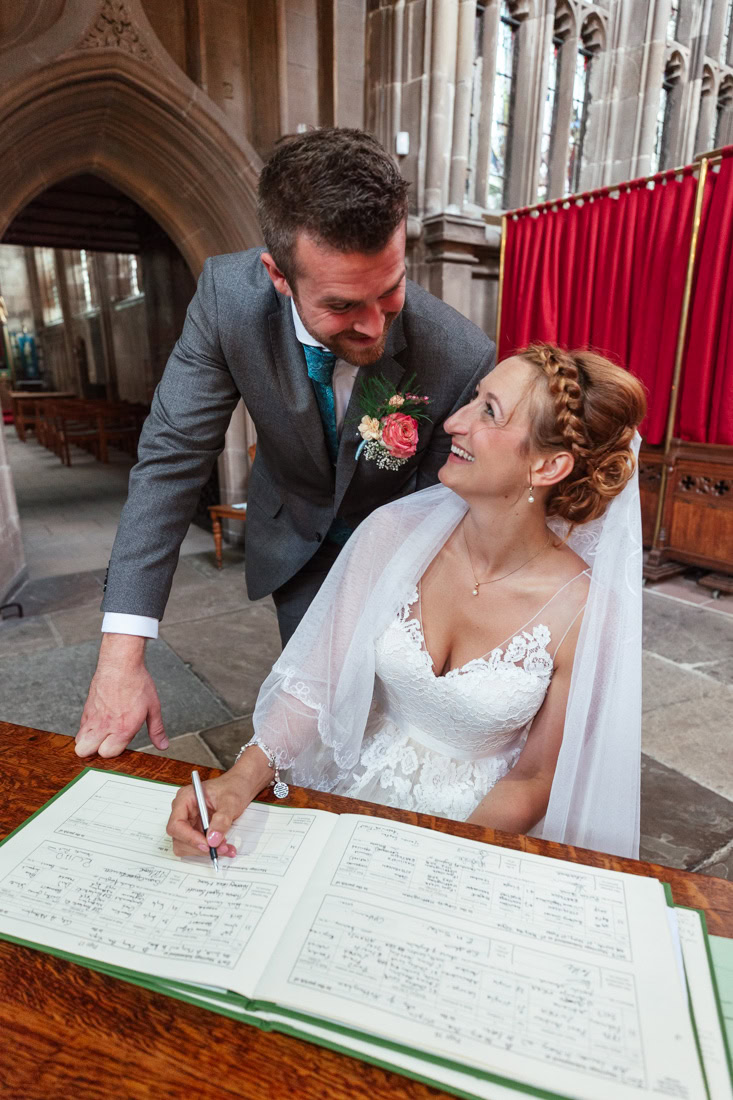 Bride signs a document in a church as groom in gray suit with floral boutonniere stands beside her.
