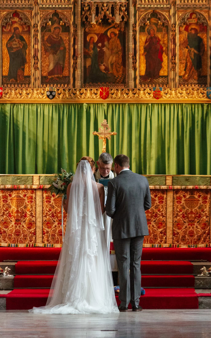 Bride and groom stand before a priest at a church altar with green curtains and religious artwork.