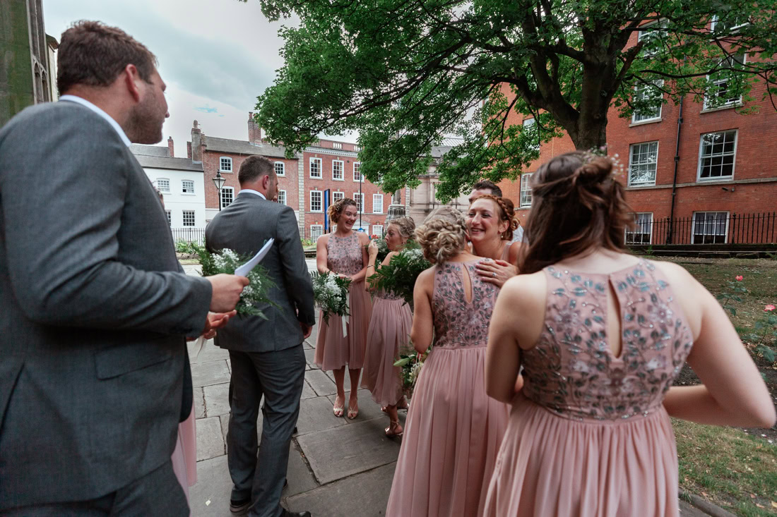 Bridesmaids in pink dresses and other guests congratulate outdoors near a tree and buildings after a wedding ceremony.