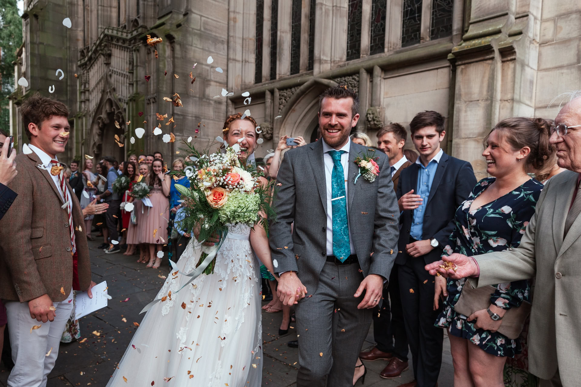 Bride and groom hold hands as guests throw confetti outside a St Mary's Church; the bride carries a flower bouquet.