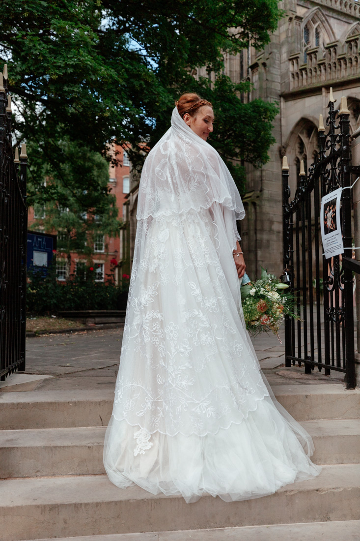 Bride in wearing a Nottingham Lace veil holds bouquet while standing on stone steps outside a gothic-style building.