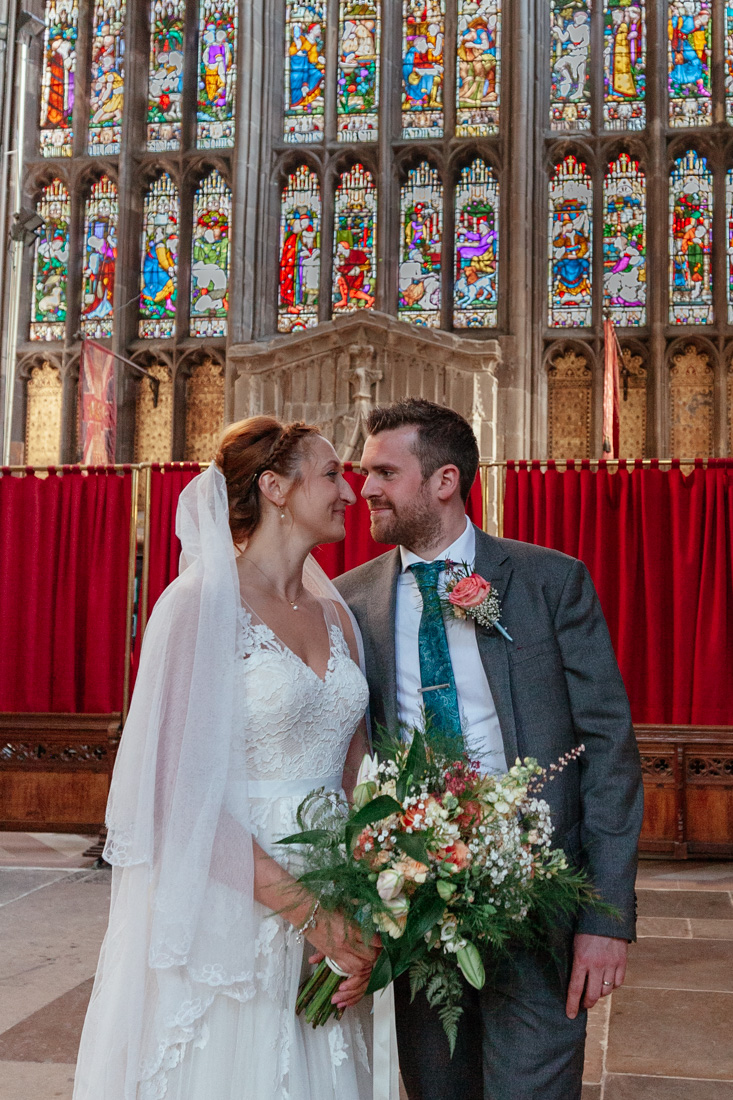 Bride and groom stand before a large stained glass window in a church; the bride holds a bouquet of flowers.