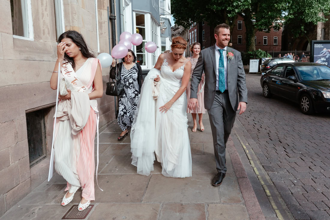 Bride and groom walk down a street together past someone wearing a “Bride To Be” sash.
