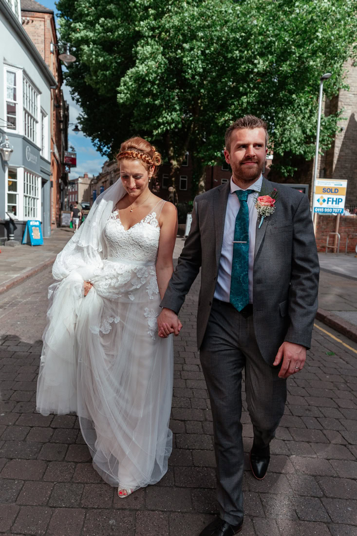 A newlywed couple walk hand in hand down a cobbled city street smiling and laughing together.