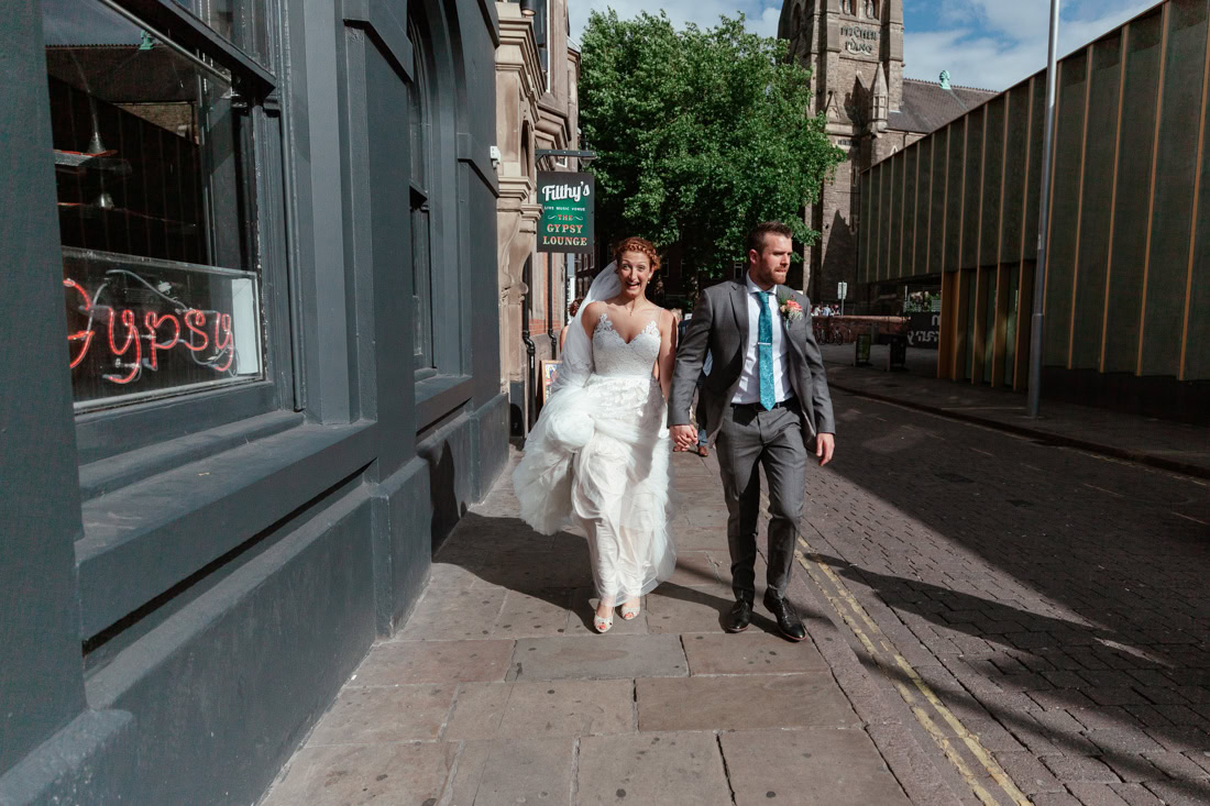 Bride in white dress and groom in gray suit walk hand-in-hand down acity street.