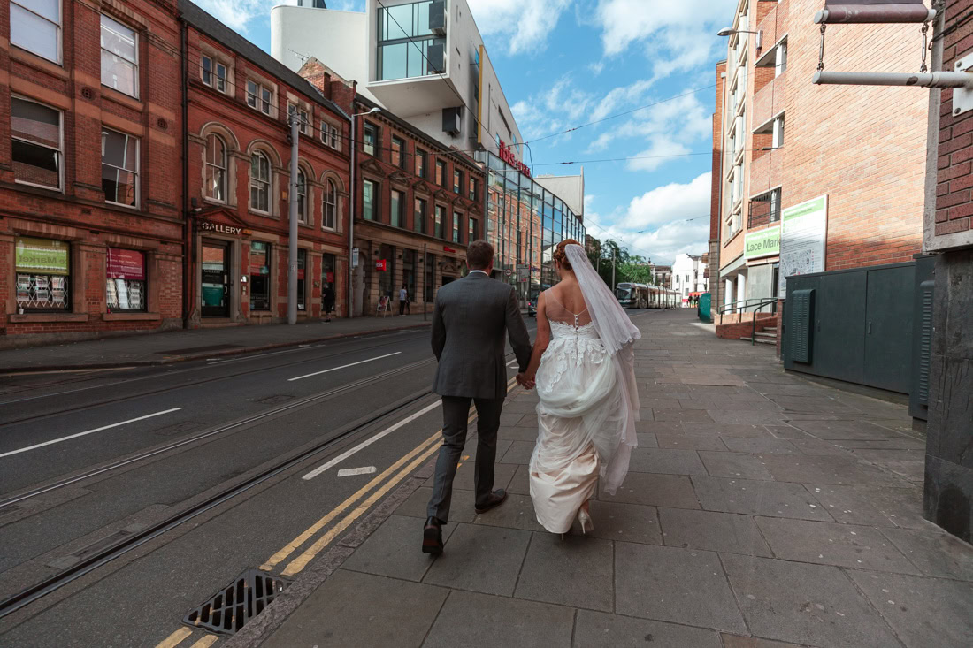 Bride and groom walk hand in hand down a city street with historic and modern buildings under a clear blue sky.