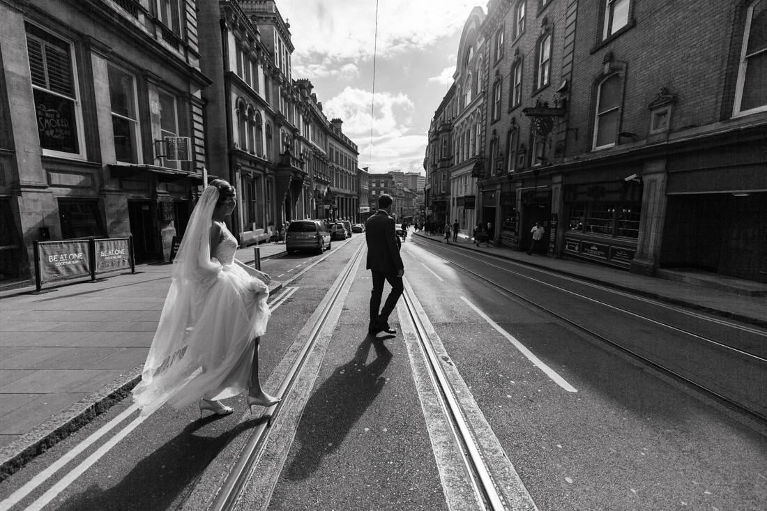 Bride and groom walk along tram tracks on a sunlit city street lined with historic buildings.