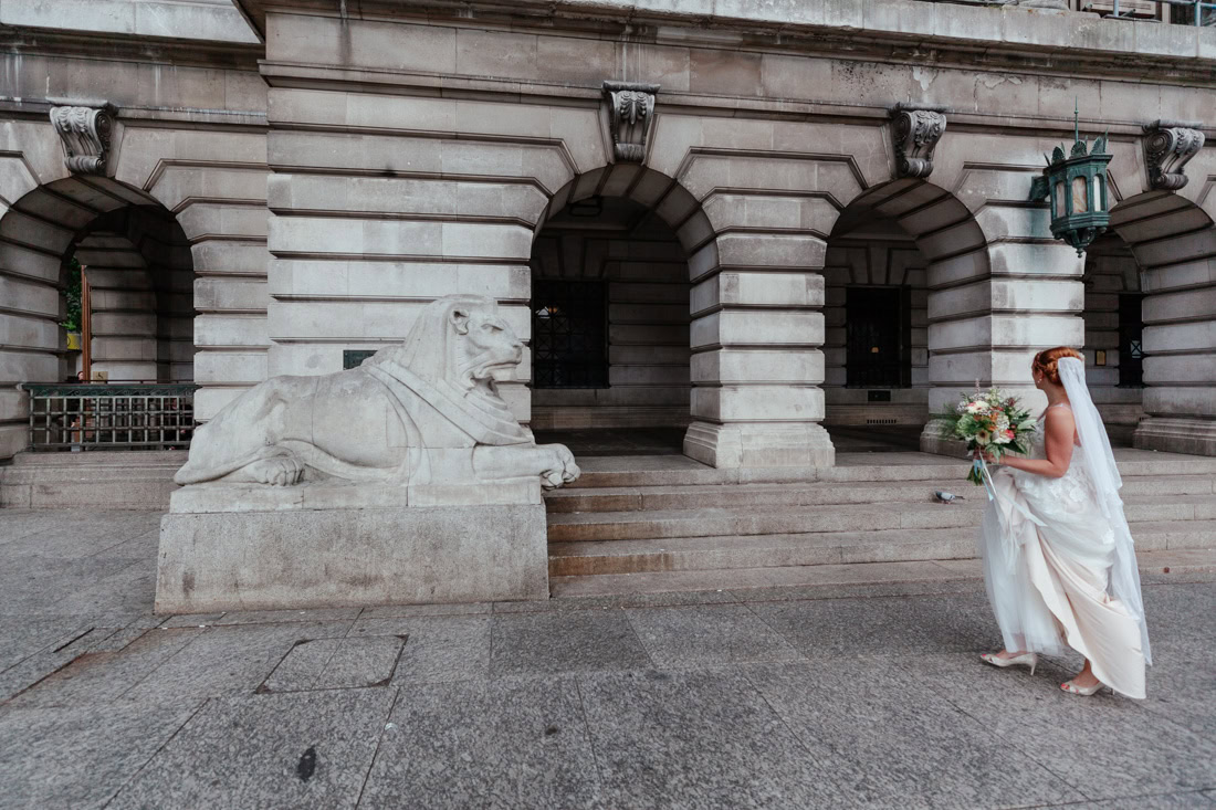 Bride holding a bouquet walks toward the Nottingham Left Lion statue outside a grand building.