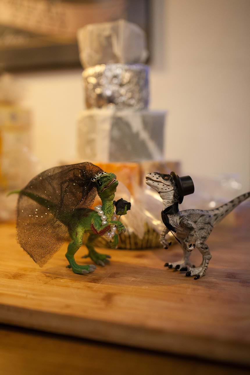 Two toy dinosaurs dressed as a bride and groom stand on a wooden surface before a tiered cheese cake.
