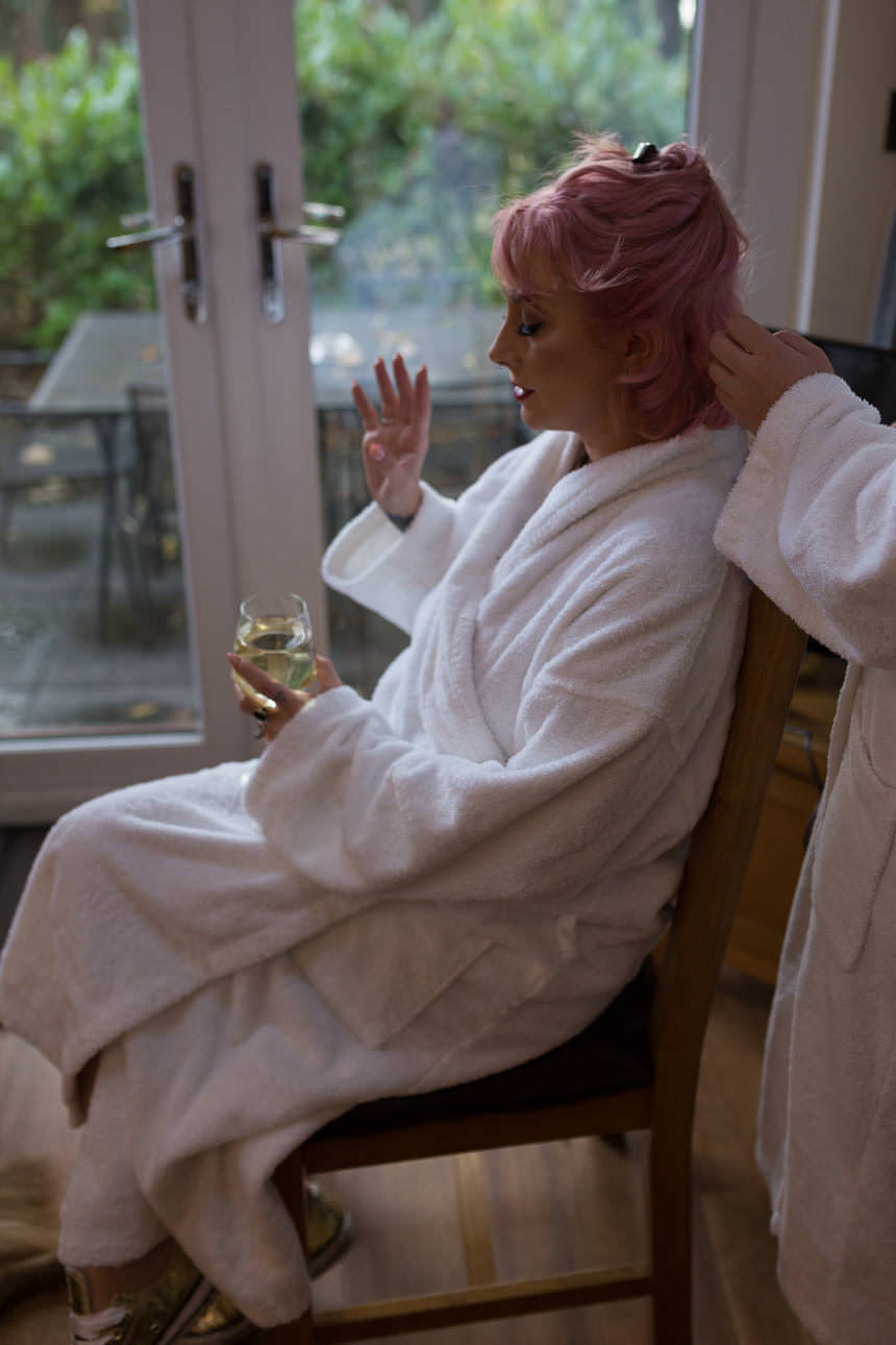 Person in a bathrobe has their pink hair styled while holding a glass of wine in a bright, cozy room.