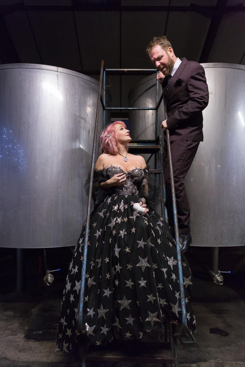 Two people in wedding attire stand on a metal staircase between large brewing tanks