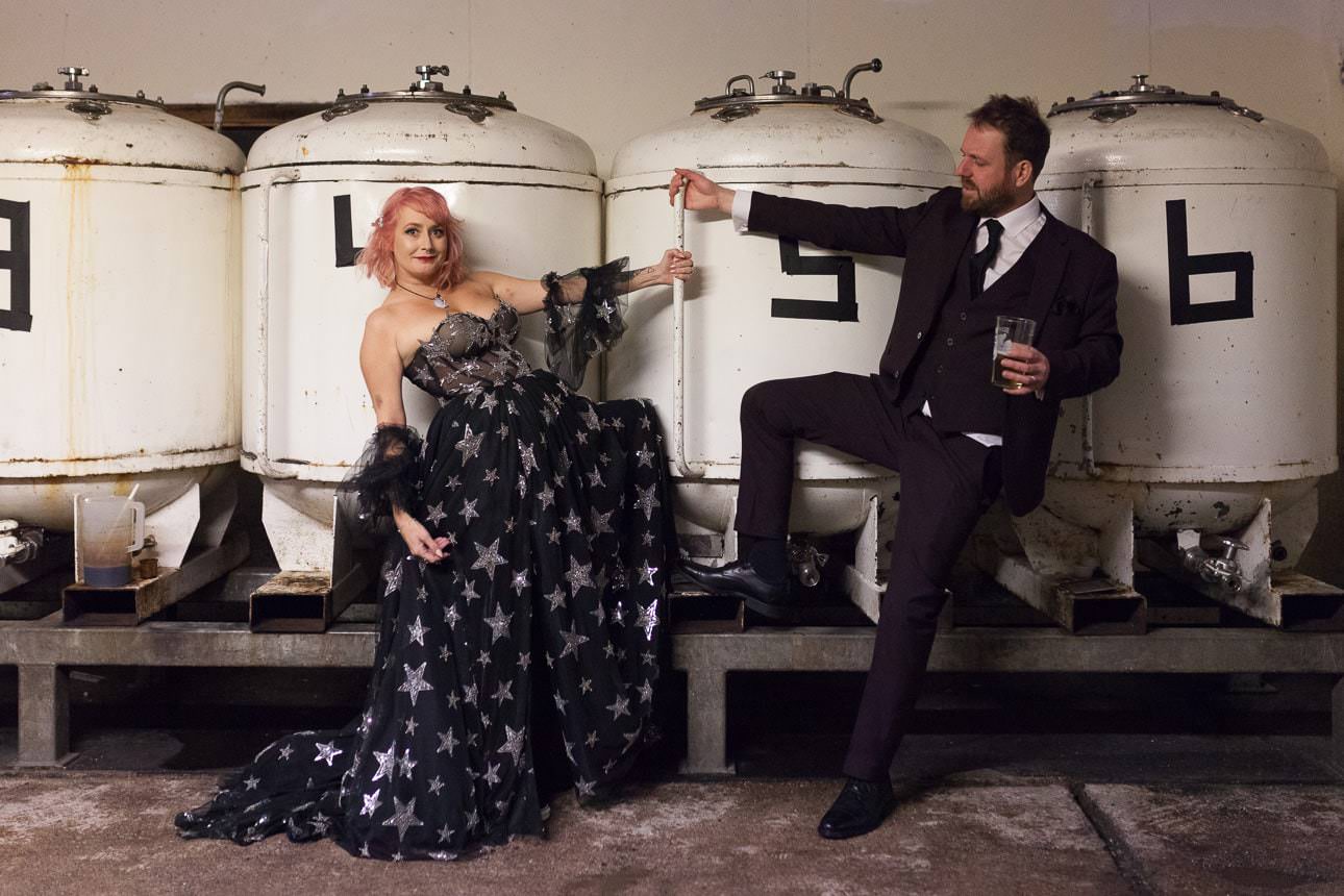 Two people in wedding attire pose playfully in front of large white brewing tanks labeled with numbers