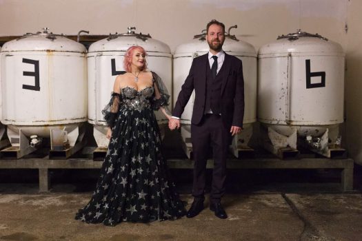 Bride and groom stand in front of brewing equipment during thier Beartown Brewery wedding.