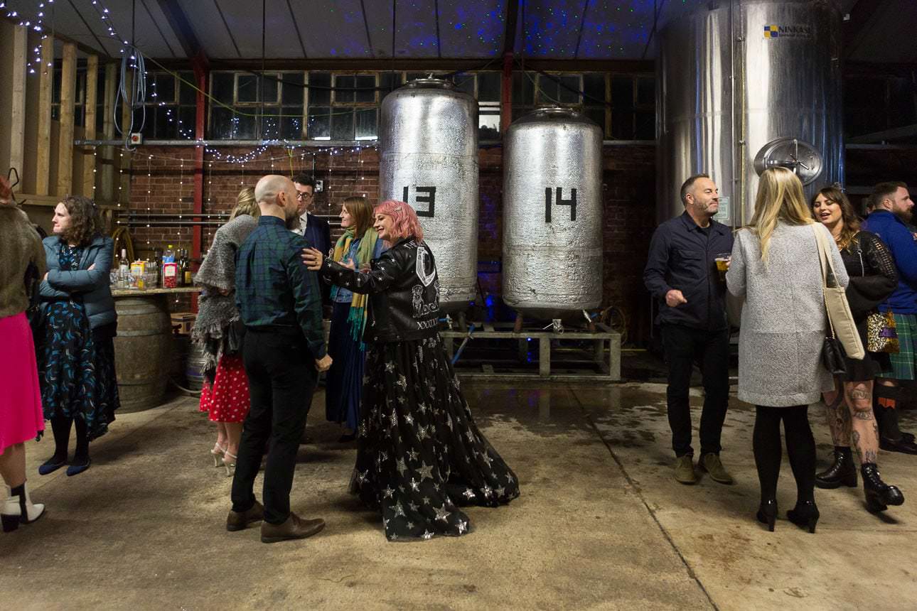 People stand and talk inside a brewery with large metal tanks and string lights overhead