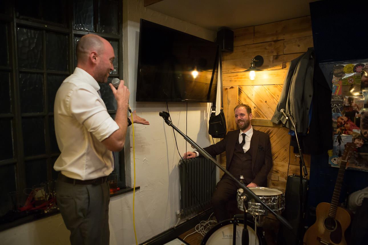 Man speaks into a microphone while another man sits smiling behind a drum set in a small room