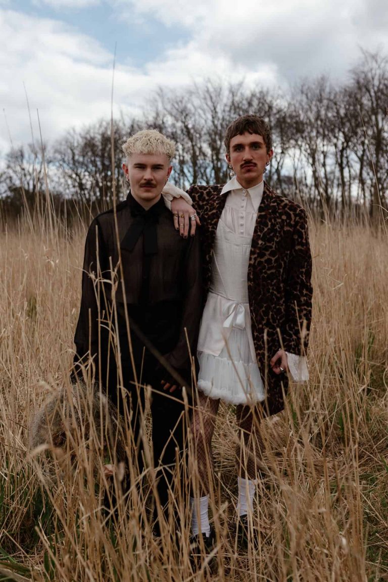 two grooms stand side by side in a field of long grass, both staring into the camera.