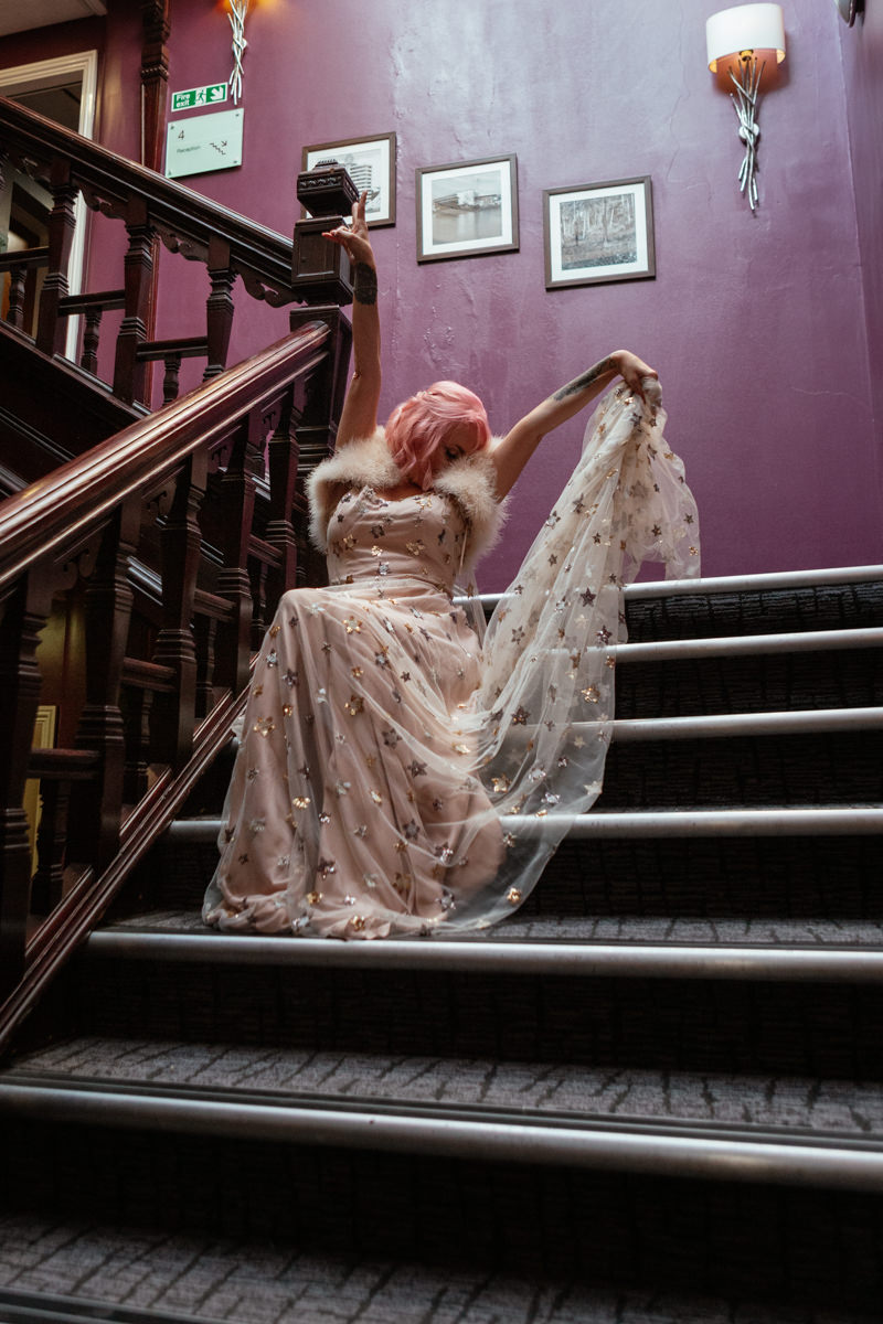 Pink-haired bride poses on purple stairwell in star-embellished gown and fur stole at micro wedding in Nottingham City.