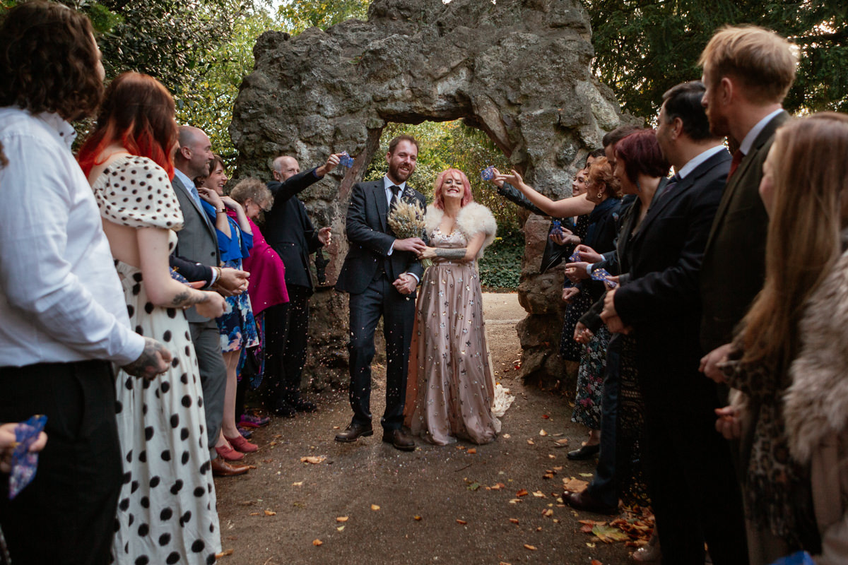 Bride and groom walk through a confetti tunnel of guests under a stone arch at Arnot Hill Park wedding.