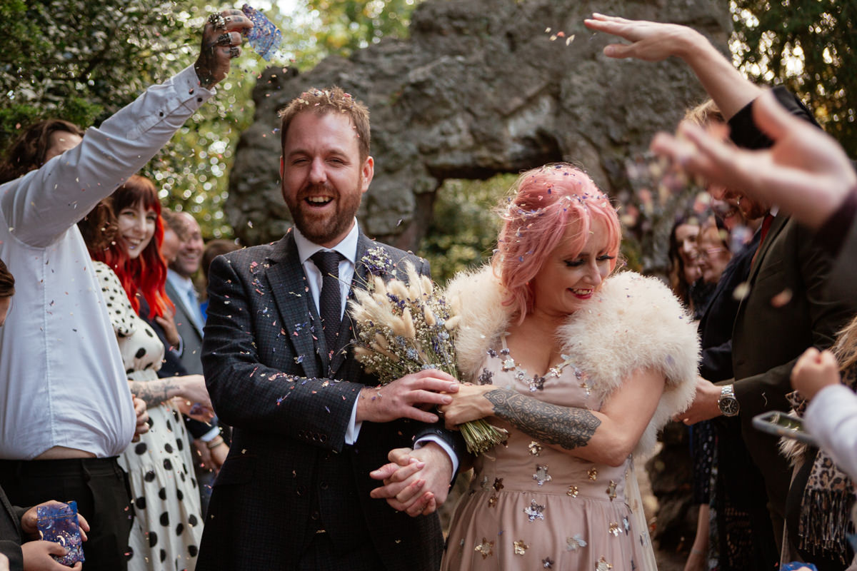 Bride with pink hair and groom laughing as guests throw confetti at Arnot Hill Park during their wedding.
