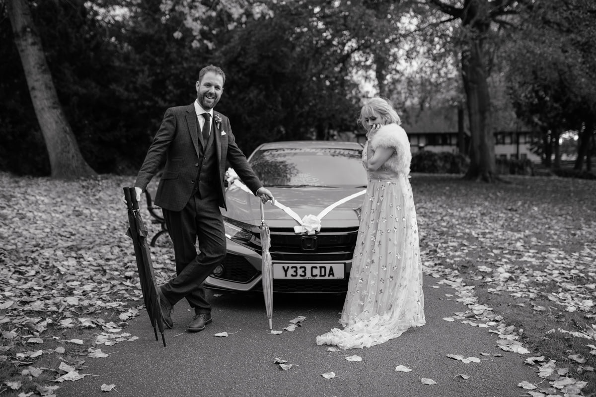 Couple in formal outfits stand before a ribbon‑decorated car on a leaf‑covered path in Arno Vale Park.