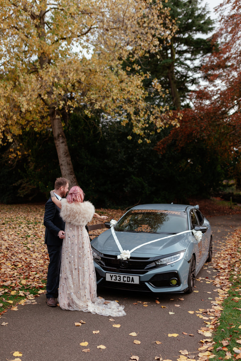 Newlyweds pose beside a decorated car on a leaf‑covered path surrounded by autumn colours.