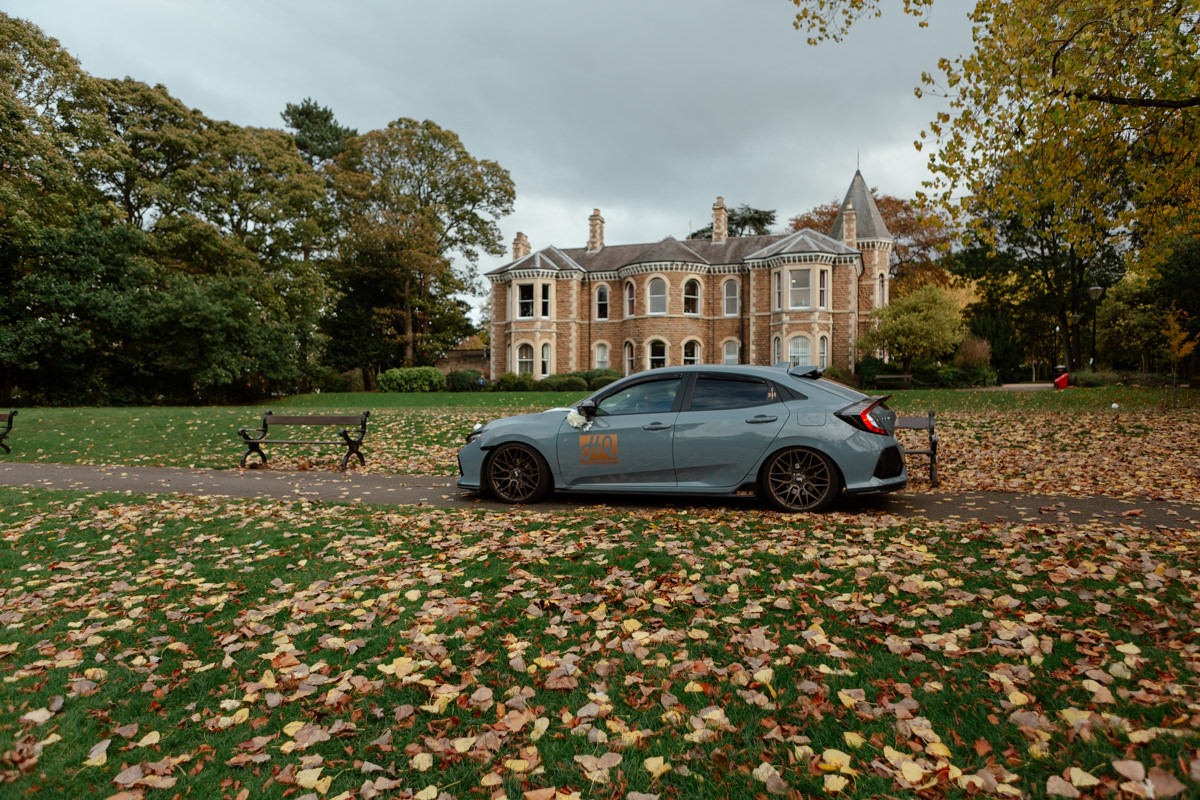 A light blue car with an “HQ” decal is parked on a leaf‑covered path in Arno Vale Park near benches and autumn trees.