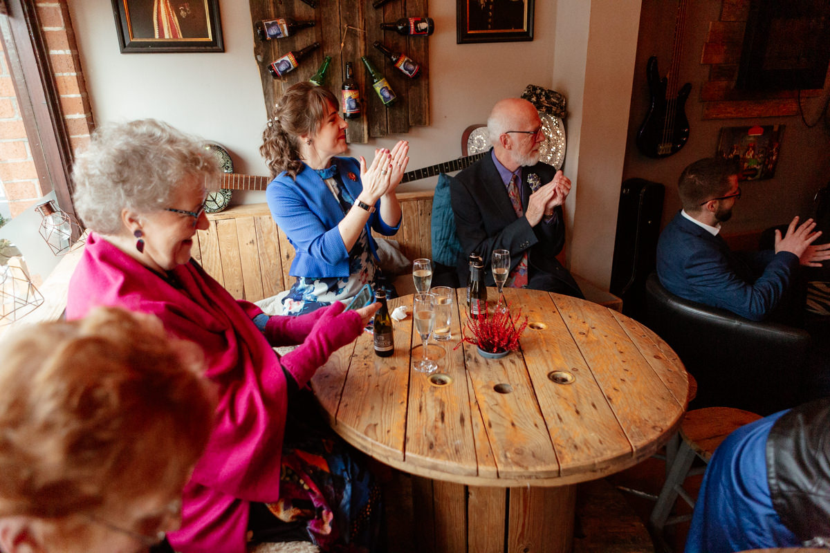A group of people sit around a wooden table clapping and enjoying drinks in a warm, rustic room.