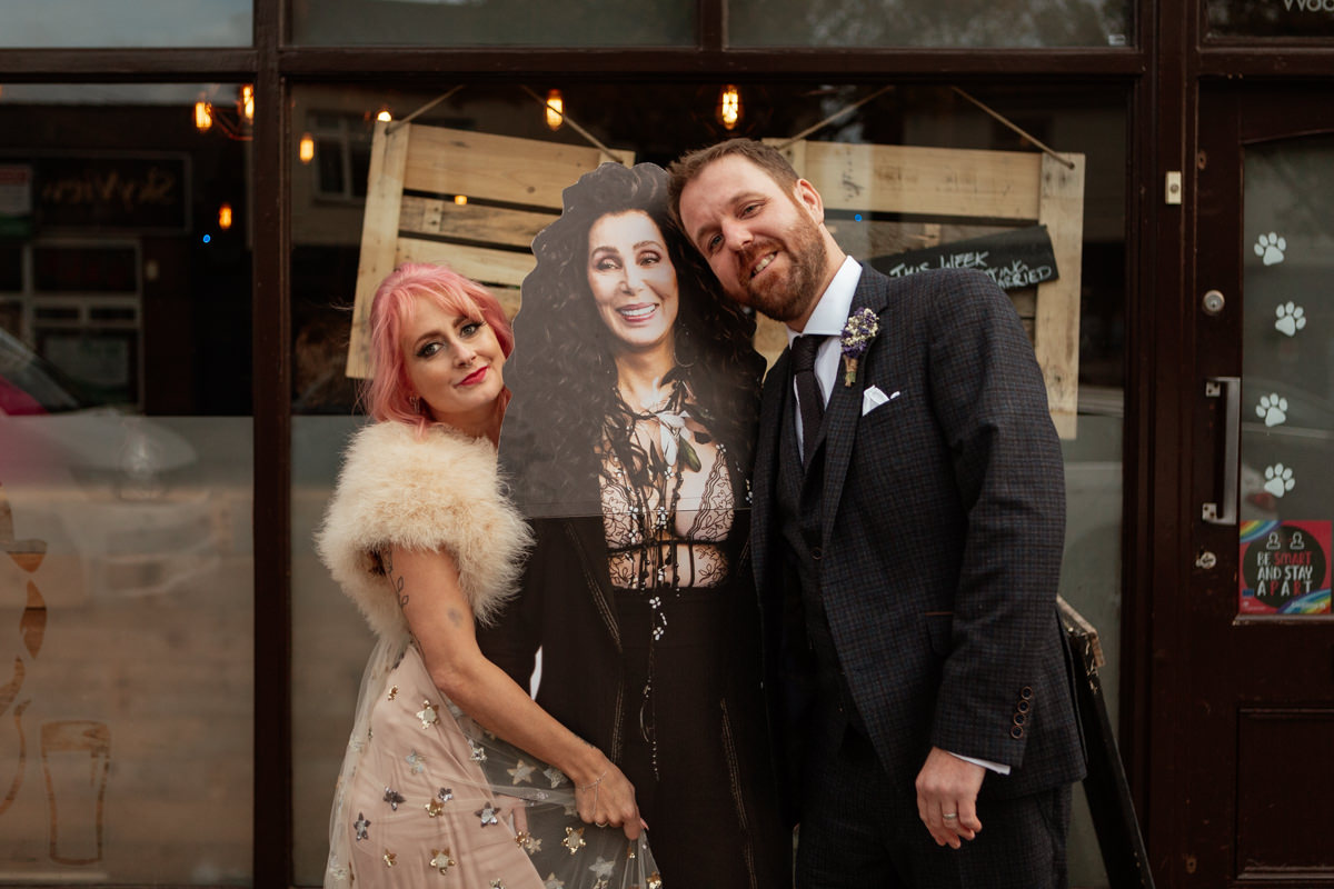 Two people in formal outfits pose outside with a life‑size Cher cardboard cutout.