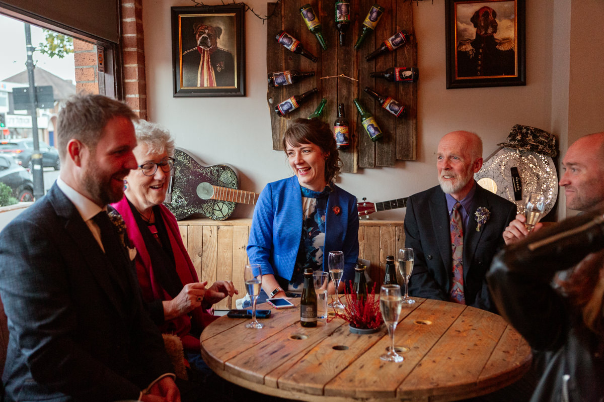 A group of people sit around a wooden table enjoying drinks in a cozy, warmly lit room.