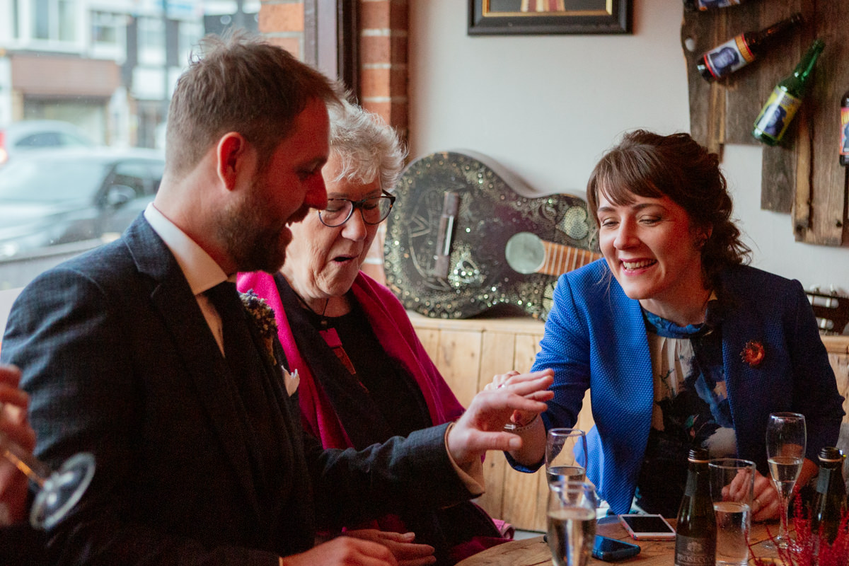 Three people sit at a wooden table, chatting as one shows his wedding ring during a celebration.
