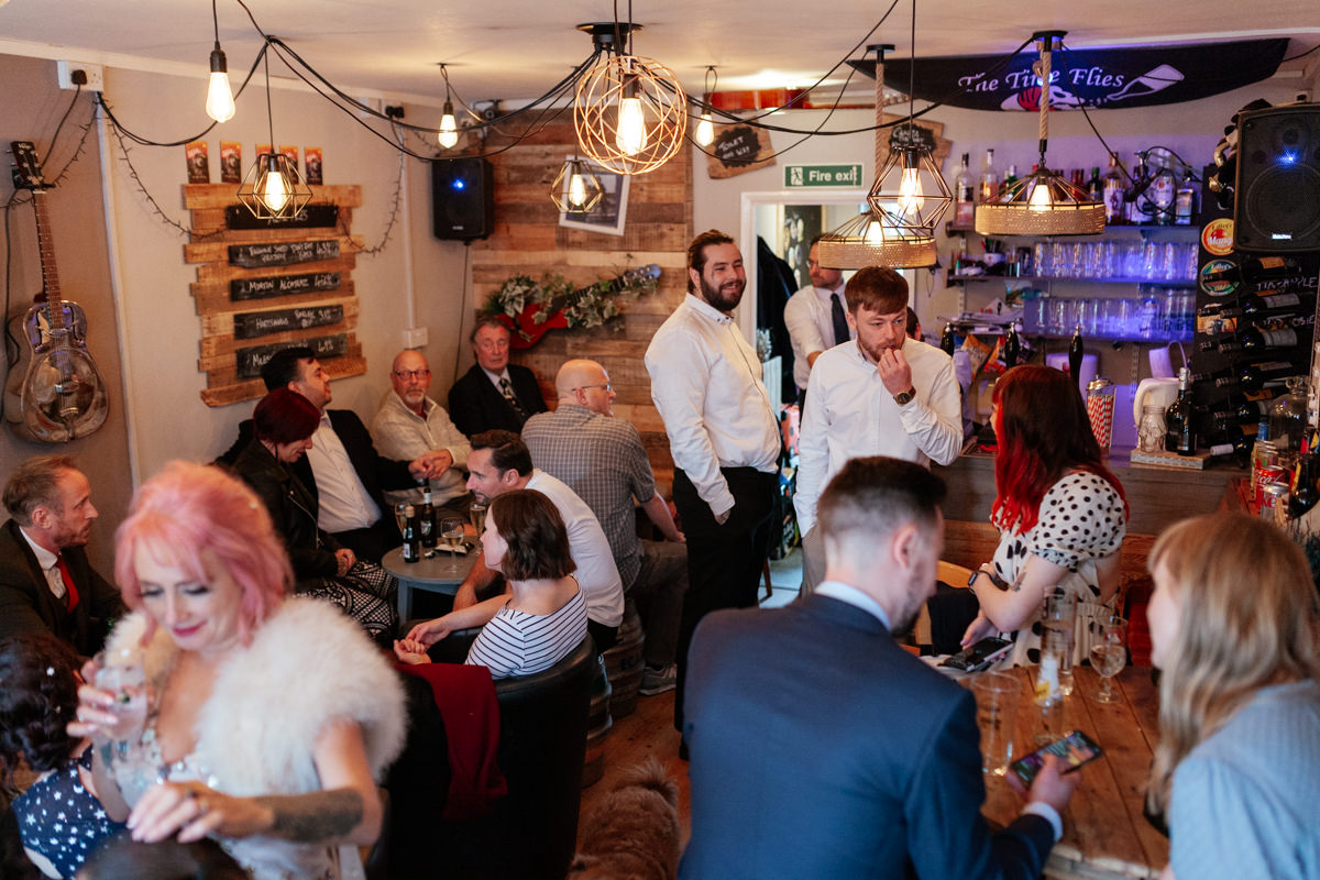 People gather in a warm, rustic bar, chatting and drinking under soft hanging lights.
