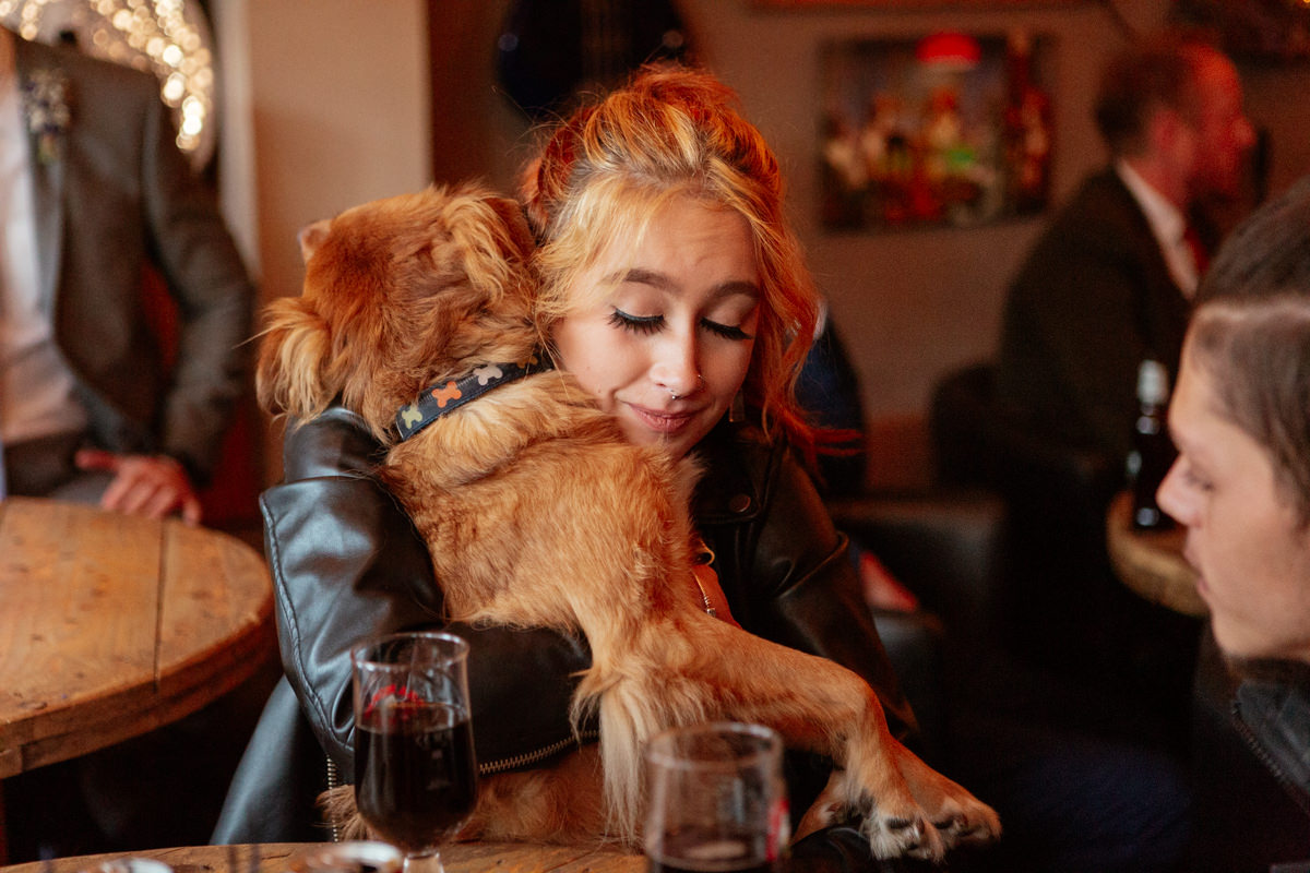 Person in a leather jacket hugs a small brown dog in a warm, busy indoor setting.