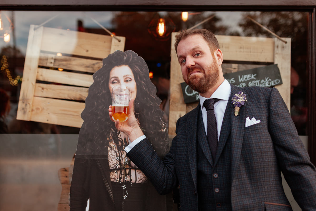 Man in a suit playfully holds a beer to a cardboard cutout of Cher's mouth.