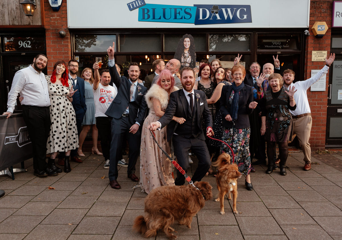 A joyful wedding party poses outside a Te Blues Dawg with two dogs at the front.